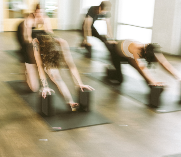Three people in a bright indoor studio stretch forward on yoga mats using yoga blocks, with motion blur emphasizing movement—capturing the dynamic energy of our Yoga Classes available in All Markets.