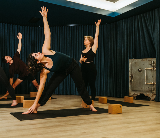 Three people practice yoga in a studio, performing a standing side stretch pose on mats while an instructor observes and guides them. Yoga blocks are placed on the floor around their mats during these Yoga Classes.