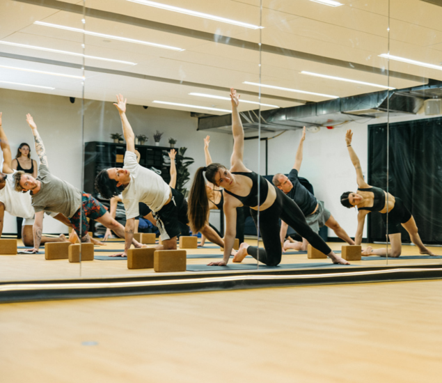 A group of people are practicing yoga in a bright, spacious studio with mirrored walls. During their Yoga Classes, they stretch sideways, one arm extended upward and the other touching a block on the floor—a welcoming space for all markets.