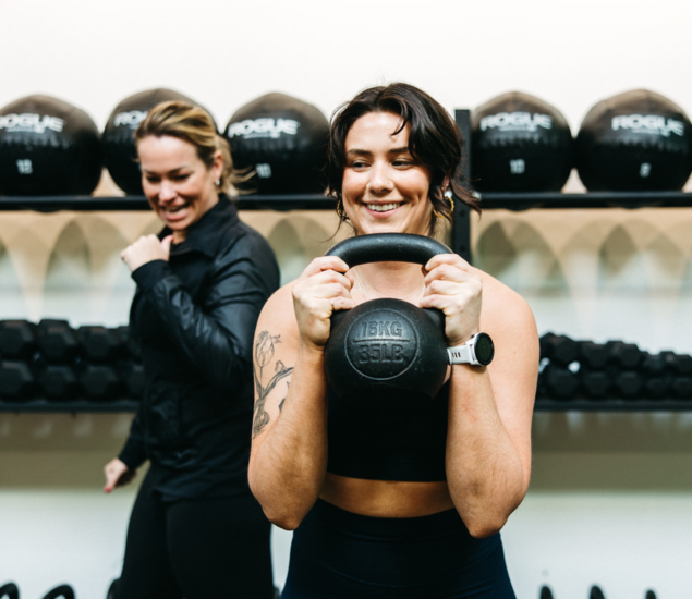 Two women at a gym smile while one lifts a 16 kg kettlebell. Rows of medicine balls and dumbbells are visible in the background. Both appear to be enjoying their fitness class together.
