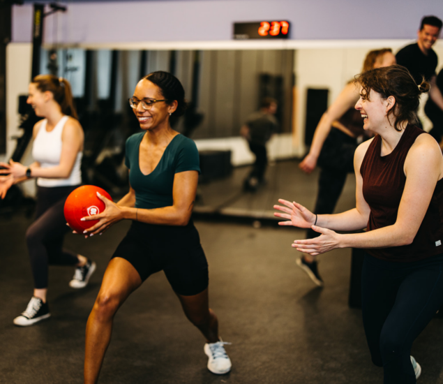 A group participates in Fitness Classes indoors. One woman holding a red ball lunges forward as others smile, clap, and encourage her. Gym equipment and a wall mirror can be seen in the background. Suitable for All Markets.