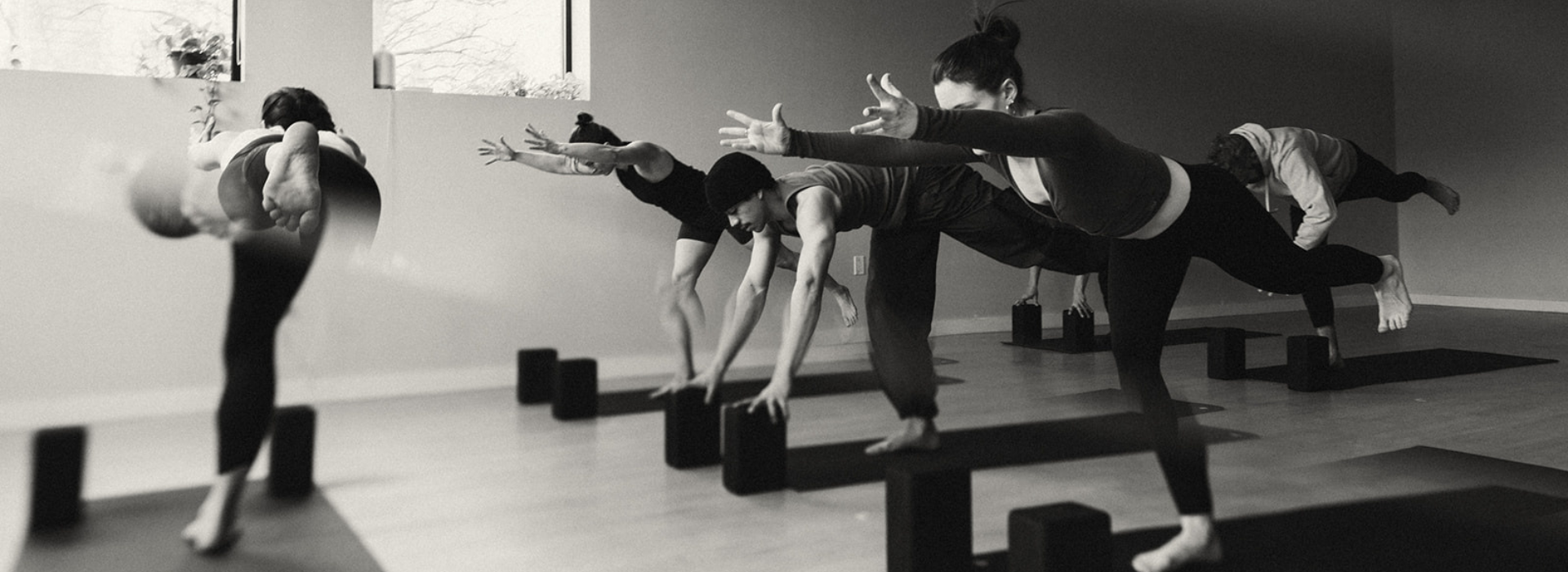 A group practices yoga in a studio, balancing on one leg with arms extended forward. Yoga blocks dot the floor, natural light streams in through high windows—captured in black and white for Summer 2026 Promos across all markets.