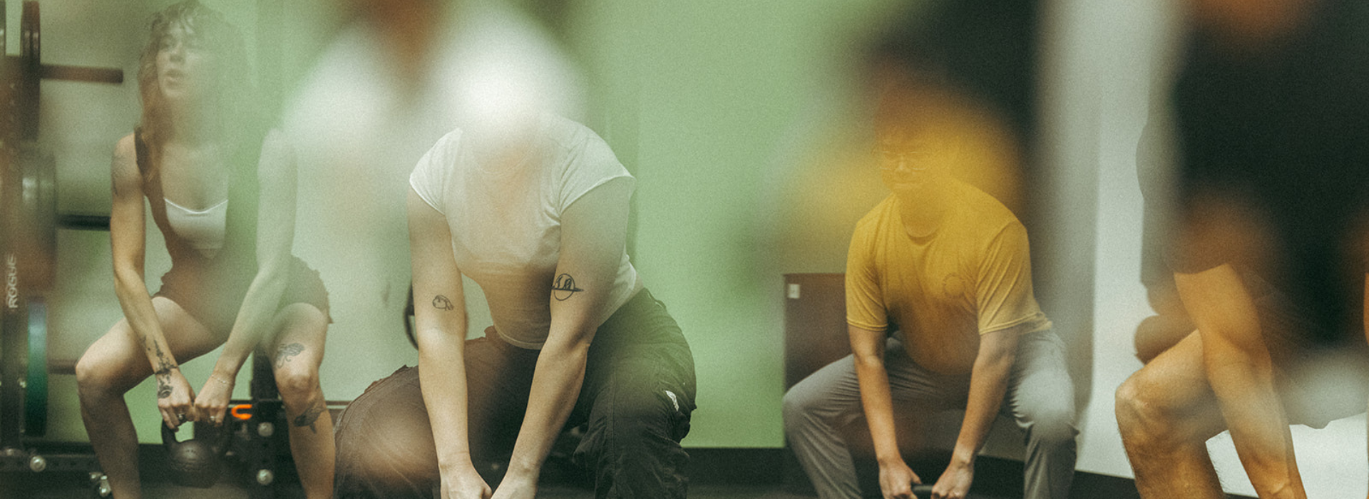 A group of people in workout attire performing squats with kettlebells during fitness classes in a studio, seen through glass with blurred reflections partially obscuring the scene.