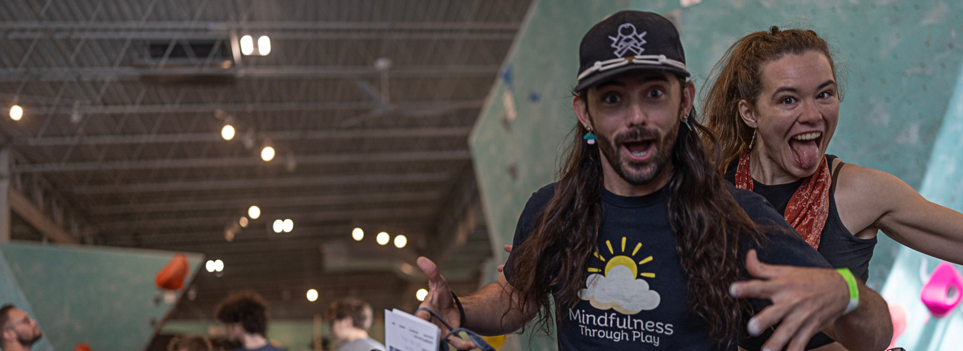 A man with long hair and a cap excitedly poses with a wide smile and open arms while a woman behind him sticks out her tongue and smiles. They are indoors at a rock climbing gym, possibly after taking climbing classes, with people in the background.
