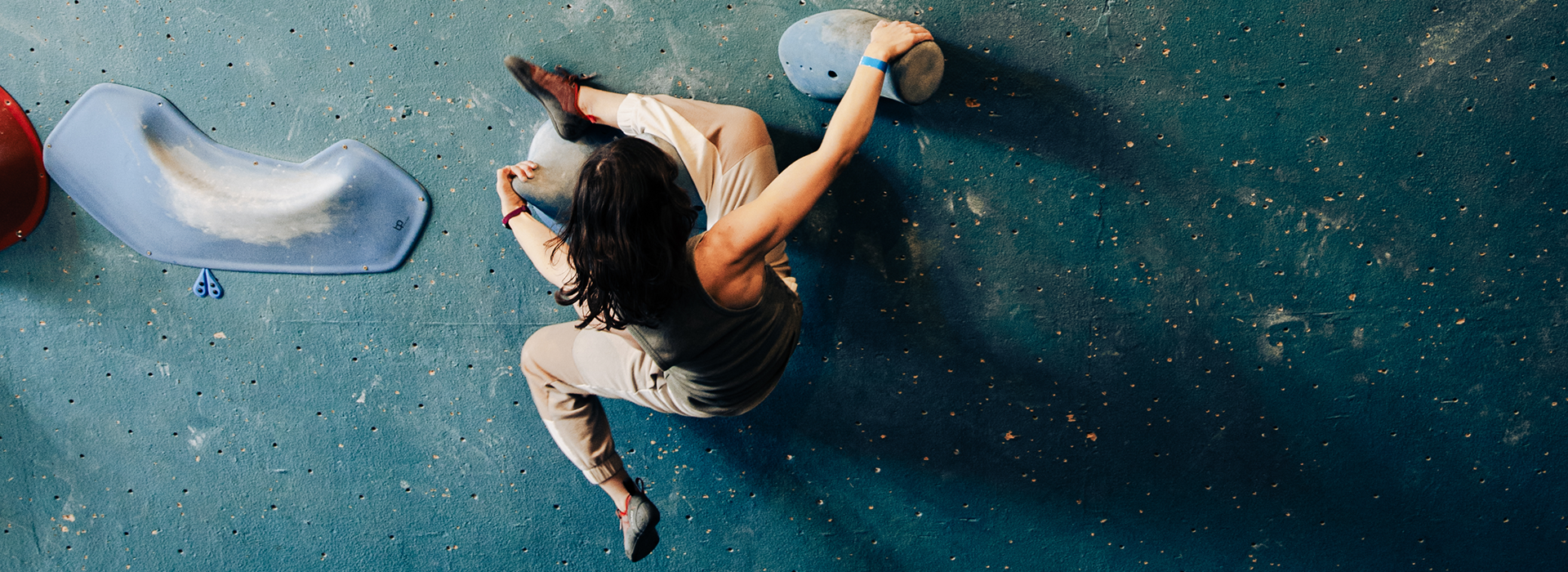 A person with long dark hair climbs an indoor bouldering wall, gripping a large blue hold with one hand and using their legs for support. The view from above showcases the textured blue wall often used in climbing classes, dotted with colorful holds.