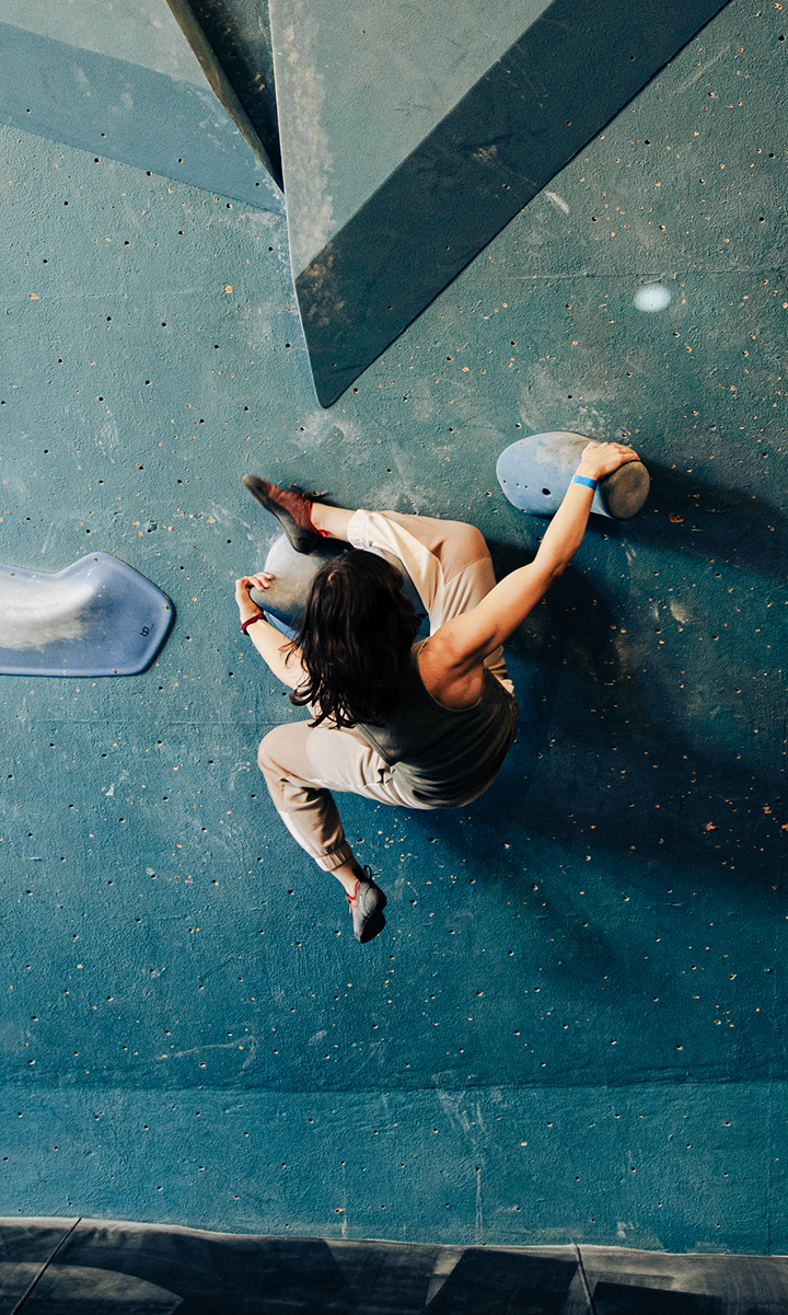 A woman with long dark hair climbs an indoor rock wall during her climbing classes, gripping large blue holds and wearing a beige top and pants, viewed from above.