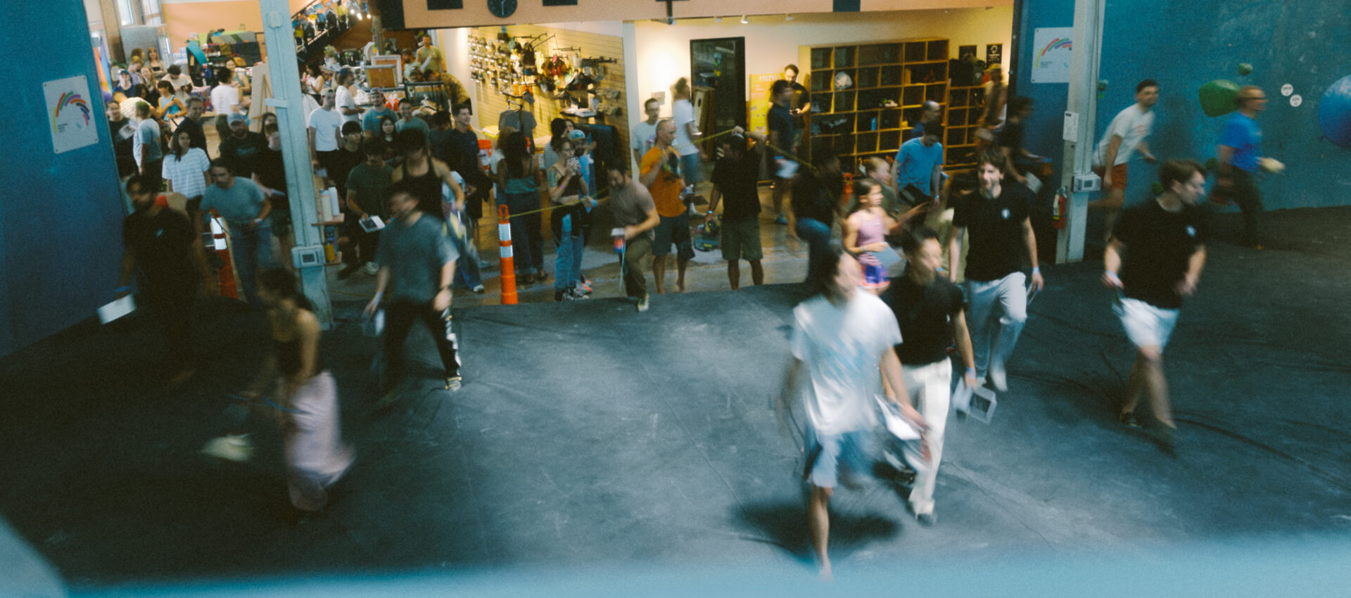 A busy indoor gym hosting lively climbing classes, with people walking and gathering—some carrying bags. On the balcony above, more spectators watch and interact, adding to the energetic atmosphere.