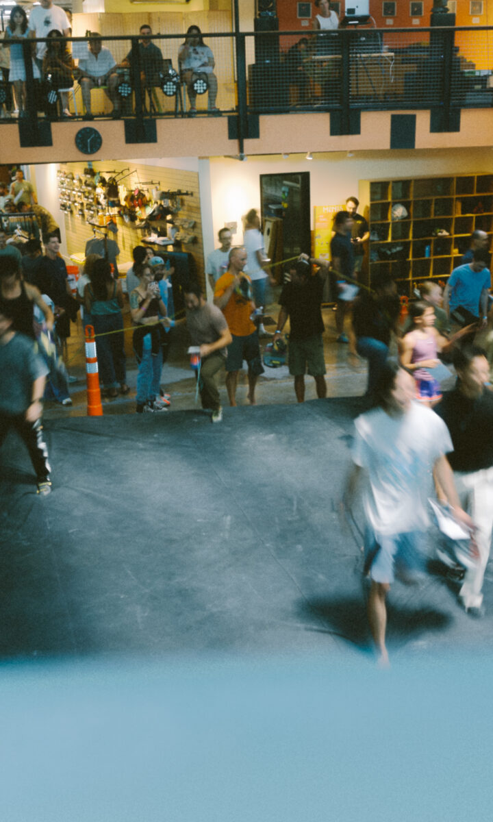 A busy indoor gym hosting lively climbing classes, with people walking and gathering—some carrying bags. On the balcony above, more spectators watch and interact, adding to the energetic atmosphere.