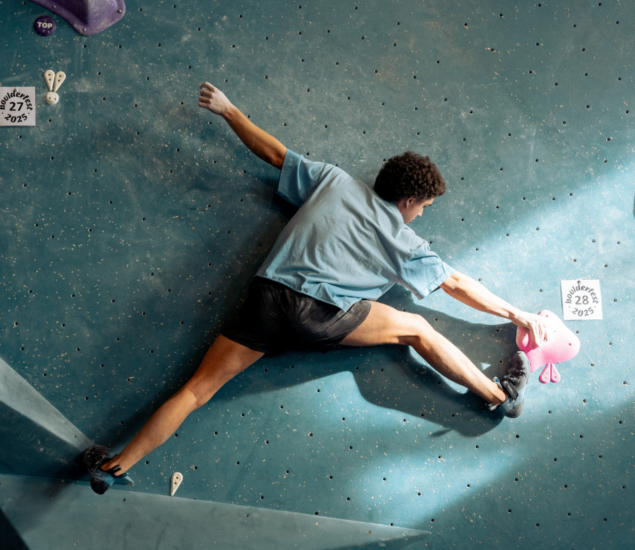 A person in a blue shirt and black shorts climbs an indoor bouldering wall, stretching wide to reach pink holds. The blue wall, featuring route labels, is a dynamic spot often used for climbing classes.