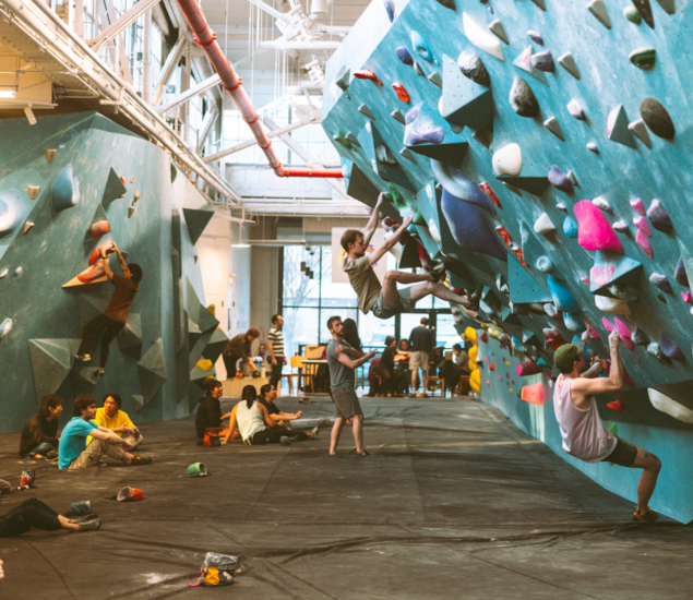 People are climbing and relaxing in an indoor bouldering gym with blue climbing walls, colorful holds, and natural light streaming in. Some attend climbing classes while others scale the walls or watch nearby.