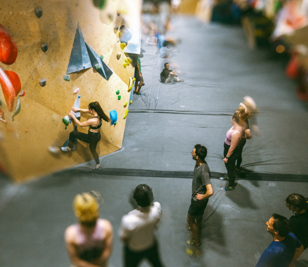 A group of people watch as a woman climbs an indoor bouldering wall with colorful holds. Some, possibly from climbing classes, stand or sit on padded flooring in the lively and energetic gym setting.