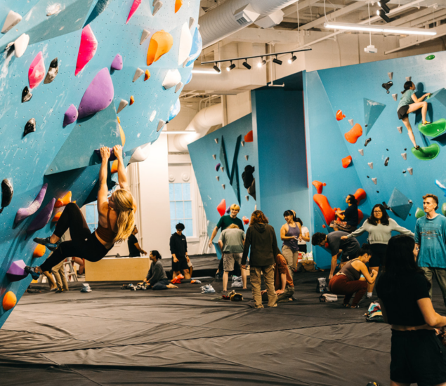 People are bouldering on colorful climbing walls in a busy indoor rock climbing gym. A woman climbs in the foreground while others, possibly taking climbing classes, climb, watch, or chat in the background on padded floors.