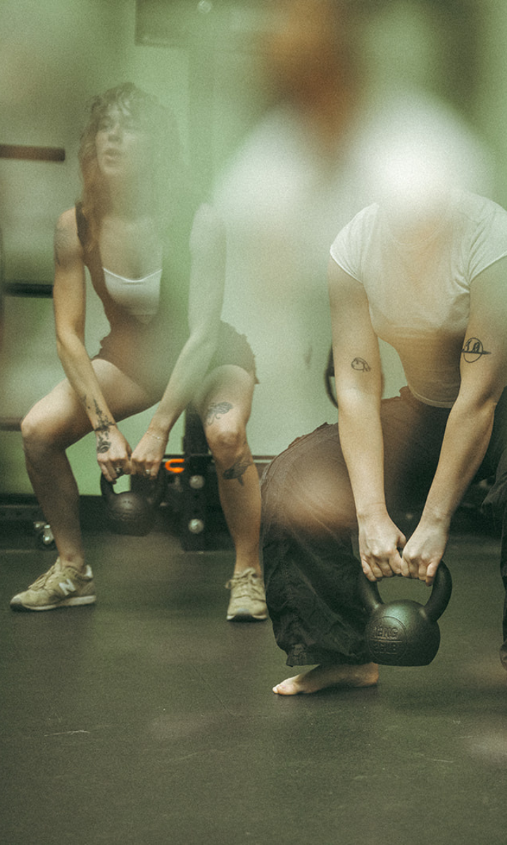 Two people exercising indoors, holding kettlebells and squatting during fitness classes. Both have tattoos on their arms and are wearing casual workout clothes. The image is slightly blurred with soft light. Suitable for all markets.