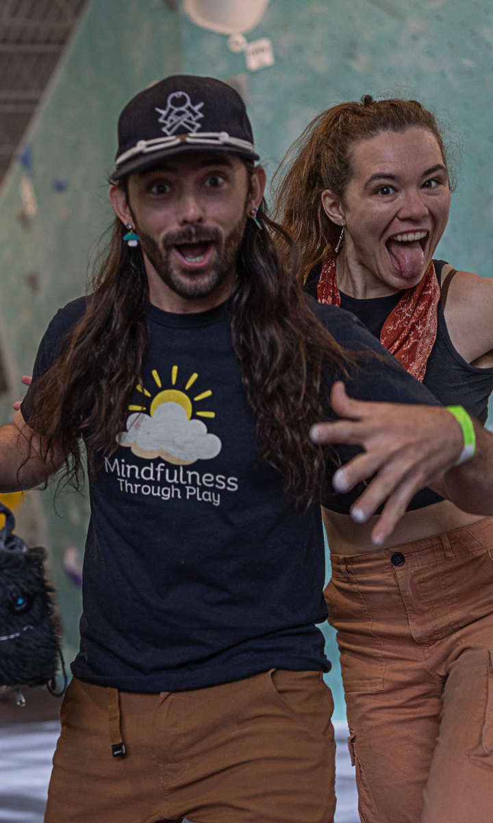 Two people in casual clothes pose playfully indoors. The man in front, wearing a Mindfulness Through Play shirt, gestures excitedly about their climbing classes, while the woman behind him smiles and sticks out her tongue. Both look happy and energetic.