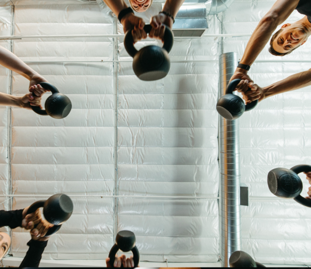 A group of people viewed from below, each holding a kettlebell with both hands, lifting them upward in a gym with white padded walls and exposed vents—capturing the energy of fitness classes offered in all markets.