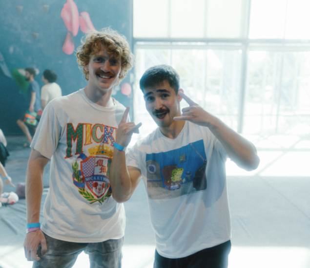 Two young men with mustaches pose indoors, smiling and making hand signs. One wears a Mickey Mouse graphic shirt, the other a SpongeBob SquarePants tee. Blurry figures and climbing holds hint at lively climbing classes in the background.