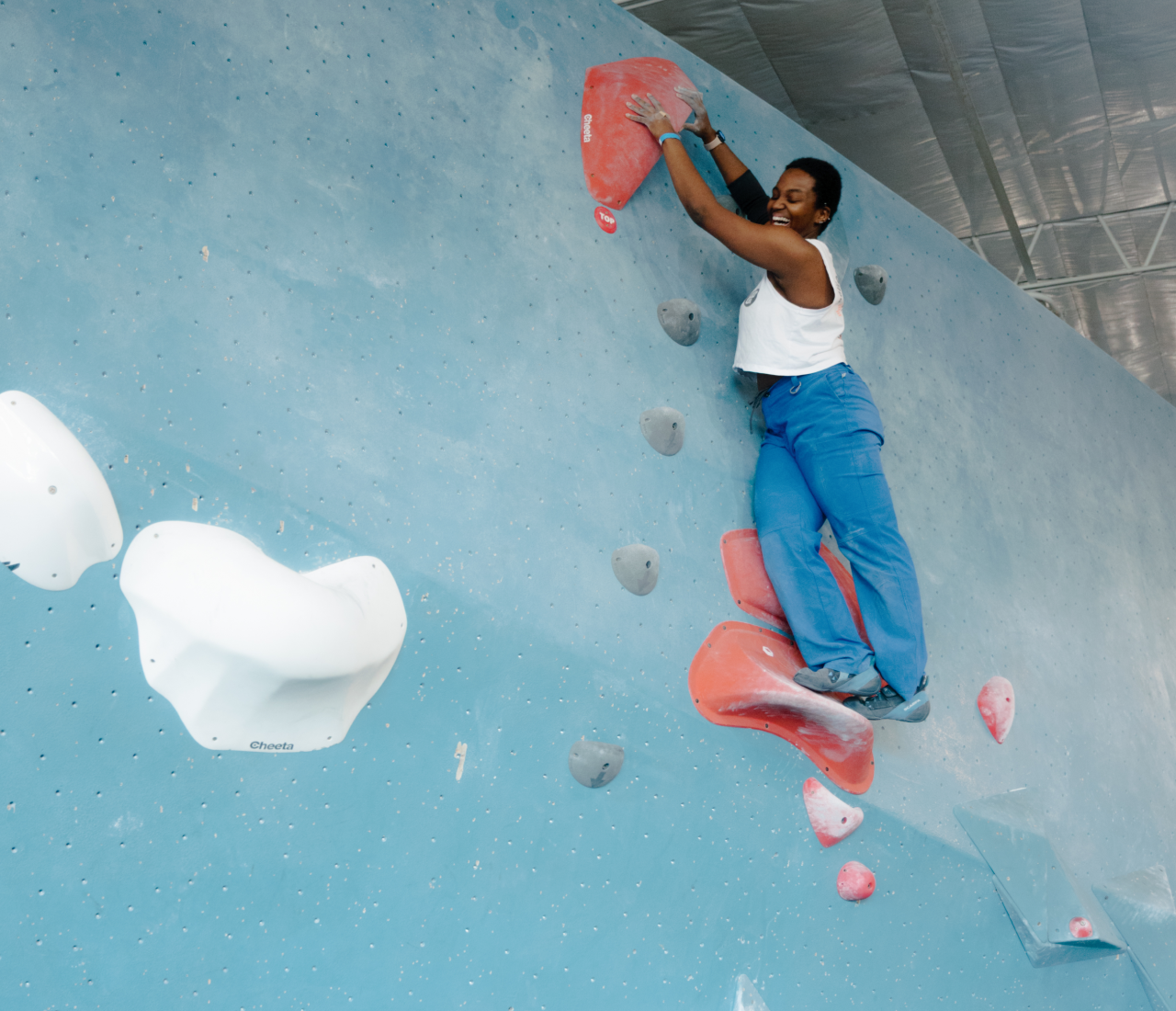 A person wearing a white tank top and blue pants climbs an indoor bouldering wall during one of the gym's popular climbing classes, gripping large red holds and smiling. The blue wall features scattered gray and white holds.