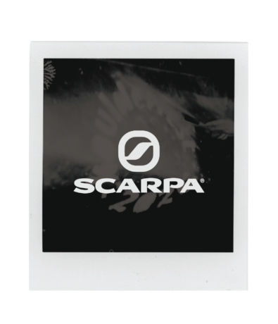 A square Polaroid-style photo features the white Scarpa logo centered on a black background, with faint patterns and shapes in the backdrop—an iconic look from Boulderfest 2026, the ultimate climbing festival.