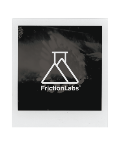 A Polaroid photo with a white border displays the FrictionLabs logo, featuring a stylized laboratory flask and mountain design above the text FrictionLabs on a dark background, captured at Boulderfest 2026, the ultimate climbing festival.