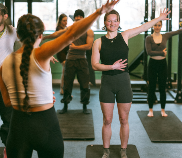 A group of people stands on exercise mats in a gym, following a fitness instructor’s movements during fitness classes. One woman in the foreground raises one arm and smiles, while others mirror her pose, showing that these sessions appeal to all markets.
