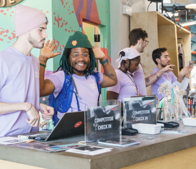 A group of four people wearing light purple shirts stand behind a counter with “Competitor Check In” signs, ready to welcome participants for climbing classes. One person in a green hat smiles and waves, while the others interact off camera.