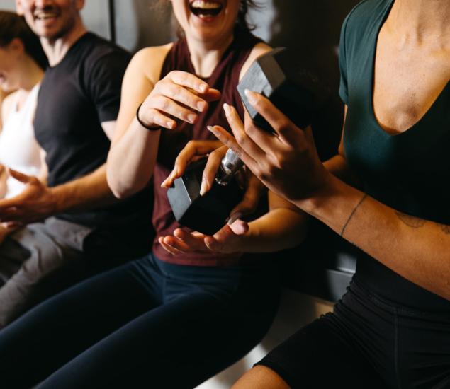 Three people in athletic wear laugh together while sitting on a bench, holding and passing dumbbells, capturing the lively energy of fitness classes available in all markets.