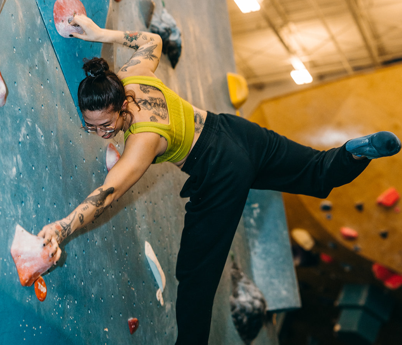 A woman with tattoos and glasses climbs an indoor bouldering wall, stretching one leg and arm wide to reach holds. She wears a yellow top, black pants, and climbing shoes—perfectly embodying the focus seen in climbing classes. Gym lighting frames more walls beyond.