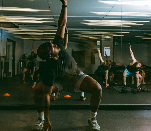 A group fitness class in All Markets stretches in front of a mirrored wall, with participants reaching one arm upward and the other touching the floor, perfectly reflecting their movements in the mirror.
