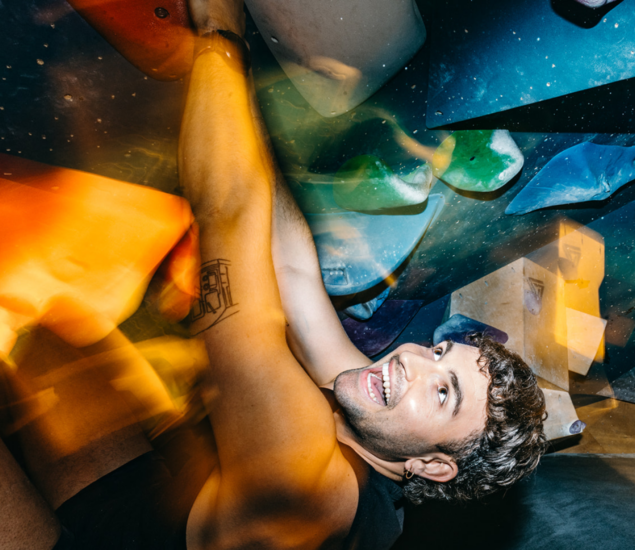 A smiling man hangs onto colorful indoor climbing holds, with a blurred motion effect creating dynamic lighting and a sense of movement—an inspiring glimpse into the excitement of climbing classes.