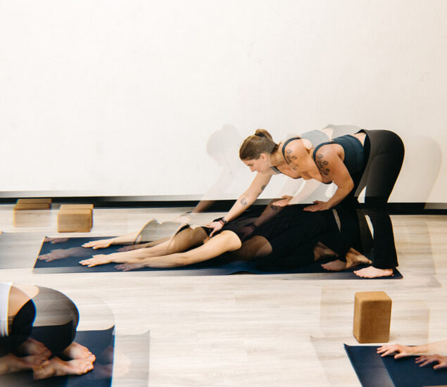A yoga instructor assists a person in child’s pose on a mat during yoga classes Brooklyn, gently pressing on their back. Yoga blocks are nearby, and other participants are partially visible against a plain white wall.