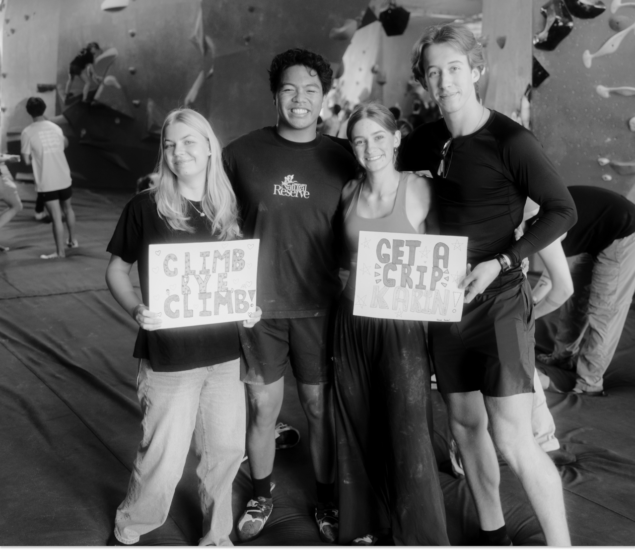 Four young adults pose and smile inside a climbing gym during the Seattle Boulderfest competition. Two hold signs: one says CLIMB CLIMB, and the other says GET A GRIP AGAIN. Climbing walls and other people appear in the background.