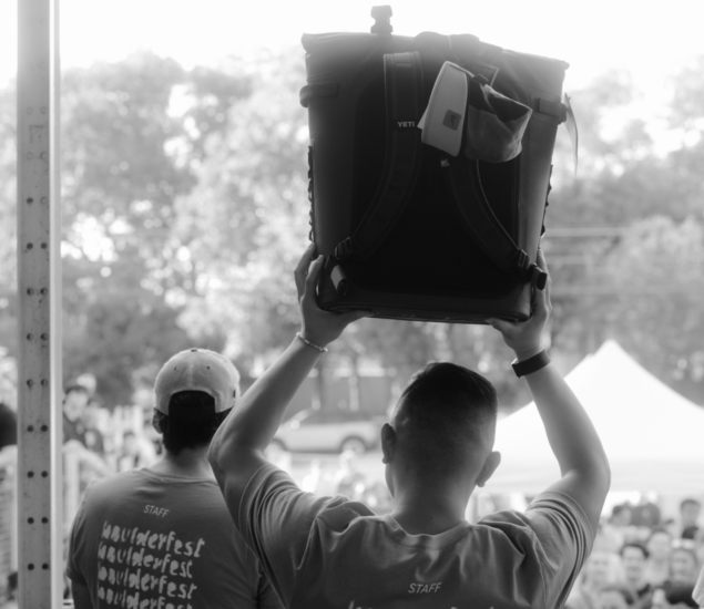 Two people in Boulderfest staff shirts face a crowd at the Seattle Boulderfest competition. One holds a large cooler above their head, with tents and trees in the background, capturing the lively outdoor event atmosphere.