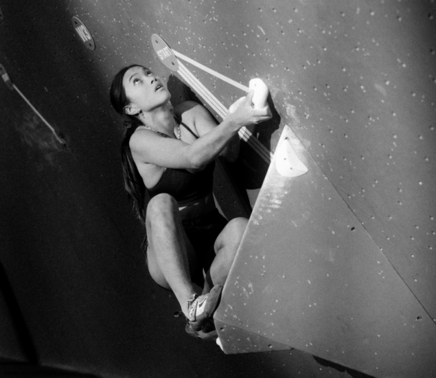 A woman climbs an indoor bouldering wall during the Seattle Boulderfest competition, gripping a handhold with both hands and pressing her foot against a triangular volume; she looks focused and determined. The image is in black and white.