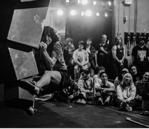 A woman climbs an indoor bouldering wall at the Seattle Boulderfest competition as a crowd watches intently; the scene is set in black and white, with dramatic lighting highlighting both the climber and the audience.