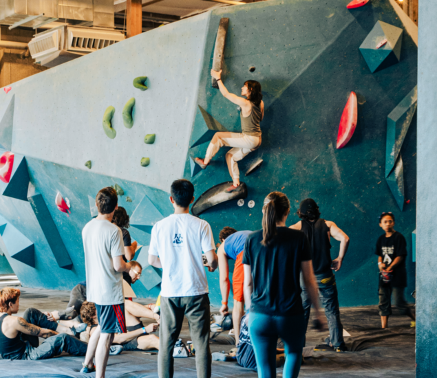 A group of people watch as a climber scales an indoor bouldering wall with colorful holds. Some, in casual athletic wear, discuss climbing classes while others sit or stand on mats in the brightly lit climbing gym.