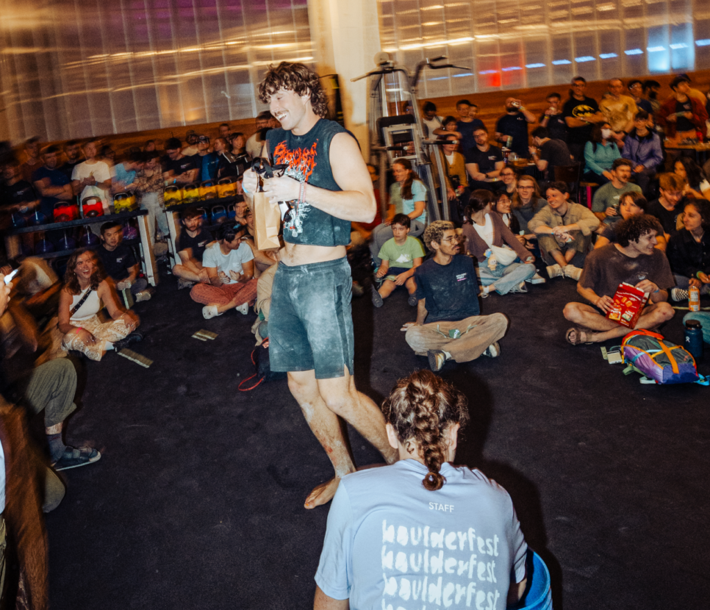A man in a sleeveless shirt stands smiling with a brown bag in a crowded indoor space, possibly after climbing classes, as people sit on the floor around him, watching and clapping. The atmosphere is lively and festive.