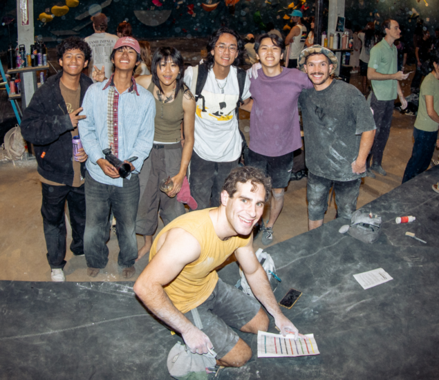A group of seven young adults pose and smile at an indoor rock climbing gym after their climbing classes; one person kneels in front holding a sheet, while the others stand behind, some with climbing gear and chalk on their clothes.