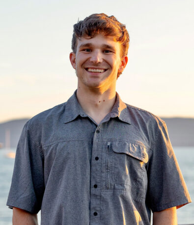 A young man with short brown hair smiles at the camera while standing outside by the water, wearing a gray short-sleeve button-up shirt. The background shows a sunset and distant hills—perfect for enjoying fitness classes Twin Cities style.