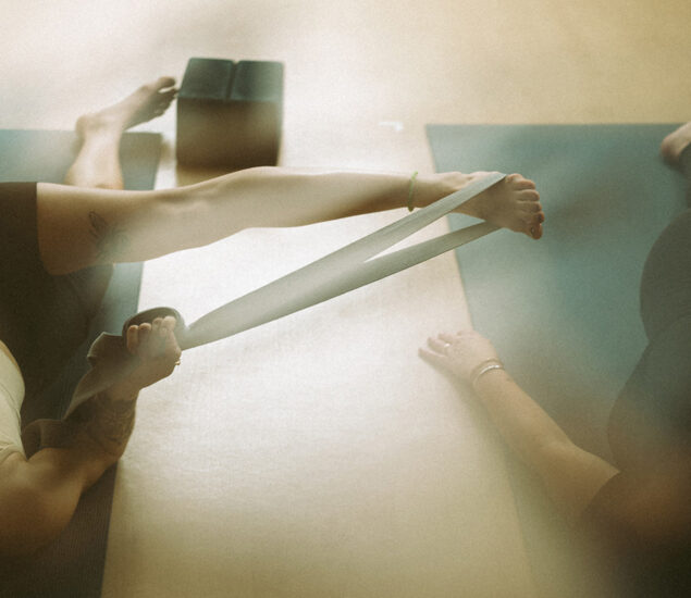 A person lying on a yoga mat stretches their leg using a resistance band, with another person nearby. A yoga block is visible in the background. Soft lighting creates a calm atmosphere, evoking the peaceful vibe of yoga classes Brooklyn offers.