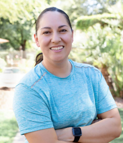 A person with dark hair pulled back, wearing a light blue athletic shirt and a smartwatch, stands outdoors with arms crossed, smiling—ready for fitness classes in the Twin Cities. Lush green trees and plants are visible in the background.