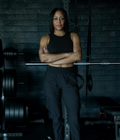 A woman in black workout clothes stands with arms crossed in a gym, facing the camera. Weightlifting equipment and a dark brick wall are visible in the background, reflecting the energetic vibe of fitness classes Twin Cities residents love.