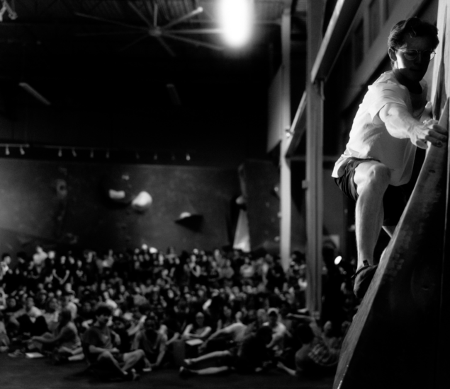 A person climbs an indoor rock wall in the foreground, with a large seated crowd watching in the background. The black and white scene appears to be a competition or event, possibly showcasing skills learned in climbing classes.