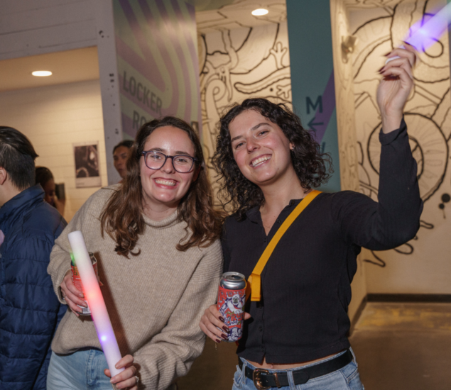 Two smiling women stand indoors at an event, one holding a glowing light stick and the other a decorated can. The background features murals and geometric designs, evoking the vibrant atmosphere of climbing classes.