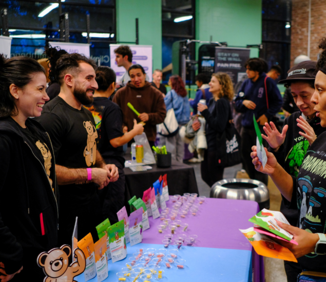 People stand at a colorful vendor booth with bear-themed snacks and lollipops, smiling and chatting about climbing classes at a lively indoor event filled with many attendees in the background.