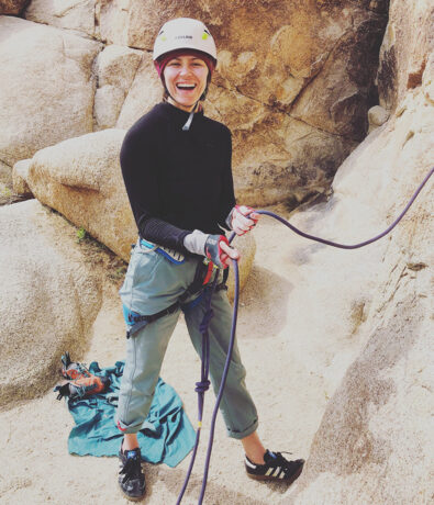 A woman wearing a helmet, gloves, and climbing gear smiles while holding a climbing rope at the base of a rocky outdoor area—her teal jacket and gear nearby—radiating the same calm confidence often seen after yoga classes in Brooklyn.