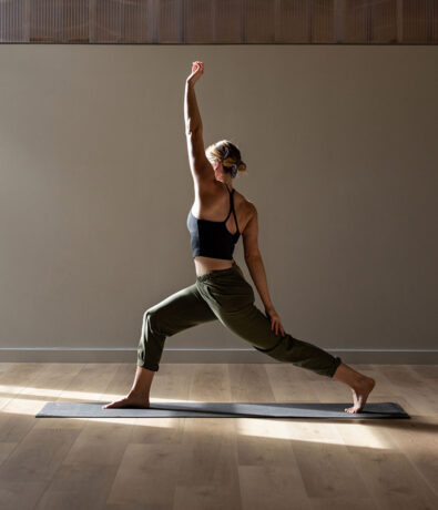 A woman practices yoga indoors on a mat, holding a warrior pose with one arm extended upward, wearing a black tank top and green pants—perfect inspiration for yoga classes Brooklyn locals will love, all bathed in natural sunlight.