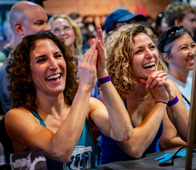 Two women with curly hair smile and clap enthusiastically in a crowded indoor setting, surrounded by others cheering after climbing classes. The atmosphere appears lively and joyful.