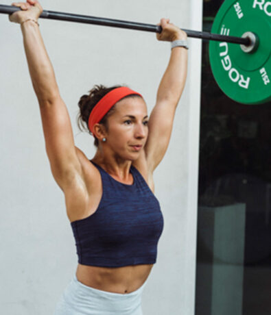 A woman wearing a red headband and navy crop top lifts a barbell with green weights overhead, standing against a plain background—her focus reflecting the intensity often seen in yoga classes Brooklyn locals love.