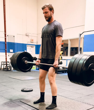 A man in a gym is lifting a heavy barbell, performing a deadlift. He wears a dark t-shirt and shorts, standing on black mats with weights and gym equipment around—typical of fitness classes in the Twin Cities.