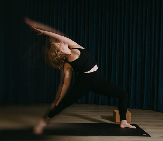 A woman in black athletic wear practices yoga on a mat indoors, performing a side angle pose with one arm reaching overhead. There is a yoga block behind her, dark curtains in the background—perfect inspiration for yoga classes Brooklyn style.